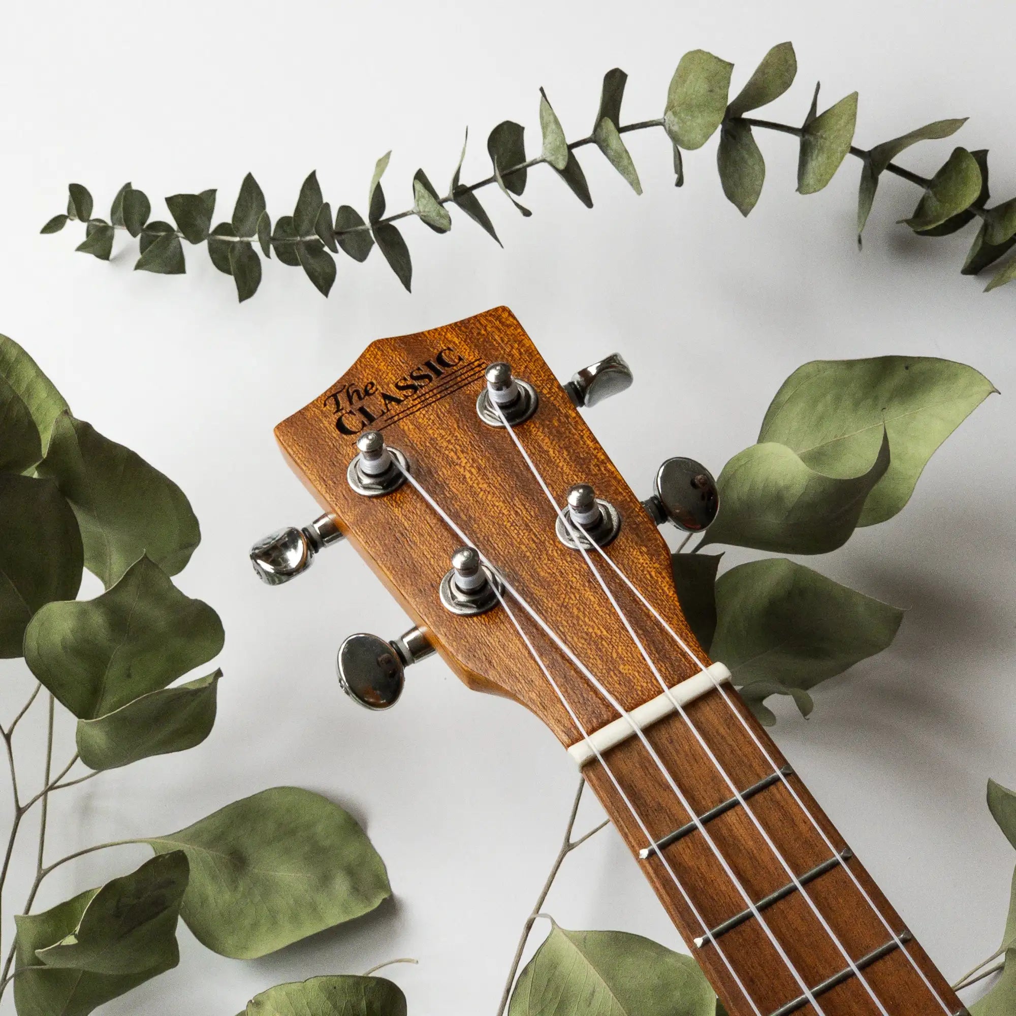 A wooden ukulele headstock with silver tuning pegs and the engraved text ’the classic’ on its surface.