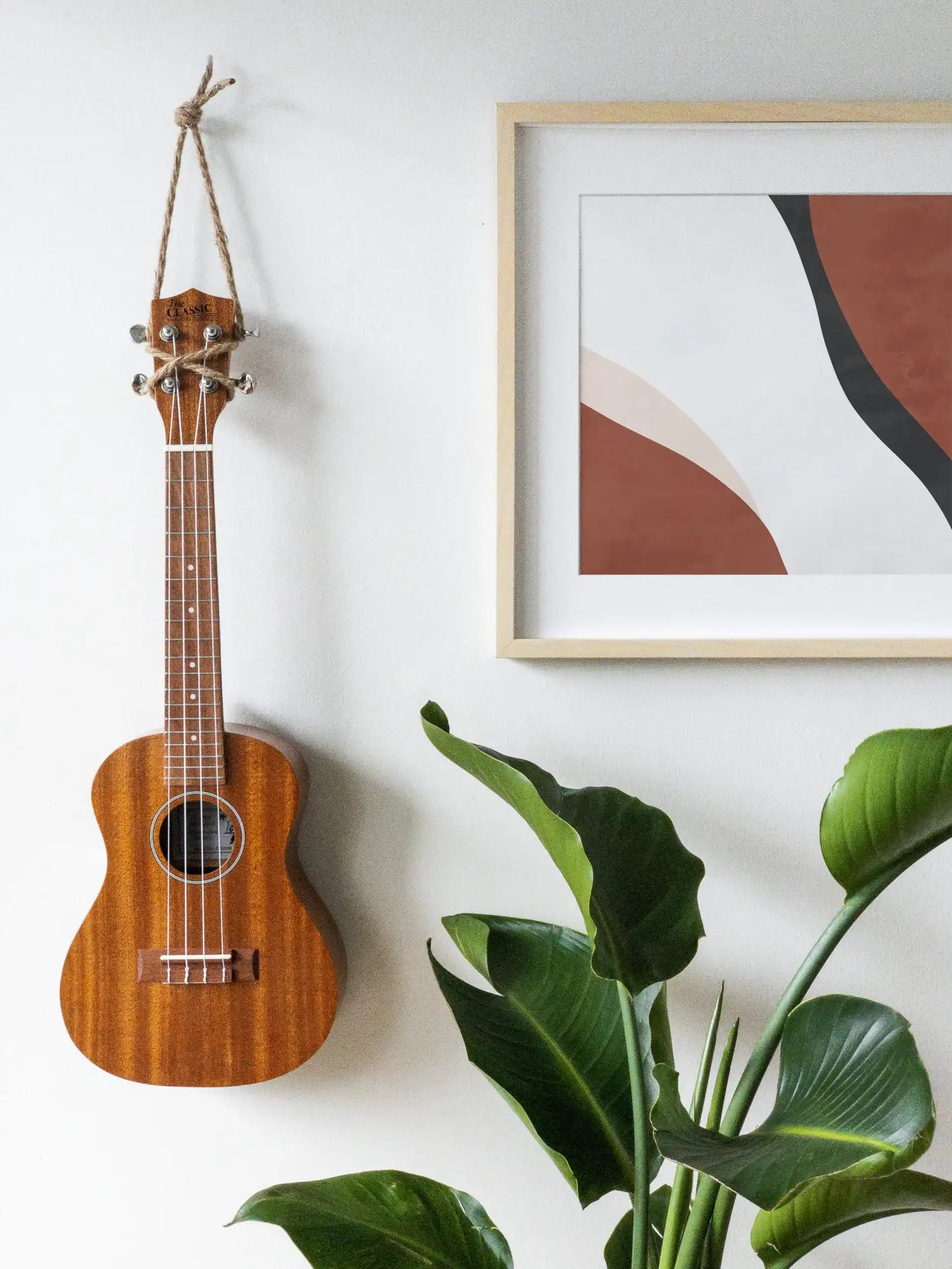 A wooden ukulele with a natural finish and silver tuning pegs hangs from a rope against a white wall.
